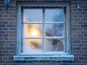 Berijpt raam van een Hollands bakstenen huis met warm goudgeel licht van binnen en koude blauwe wintermist buiten.