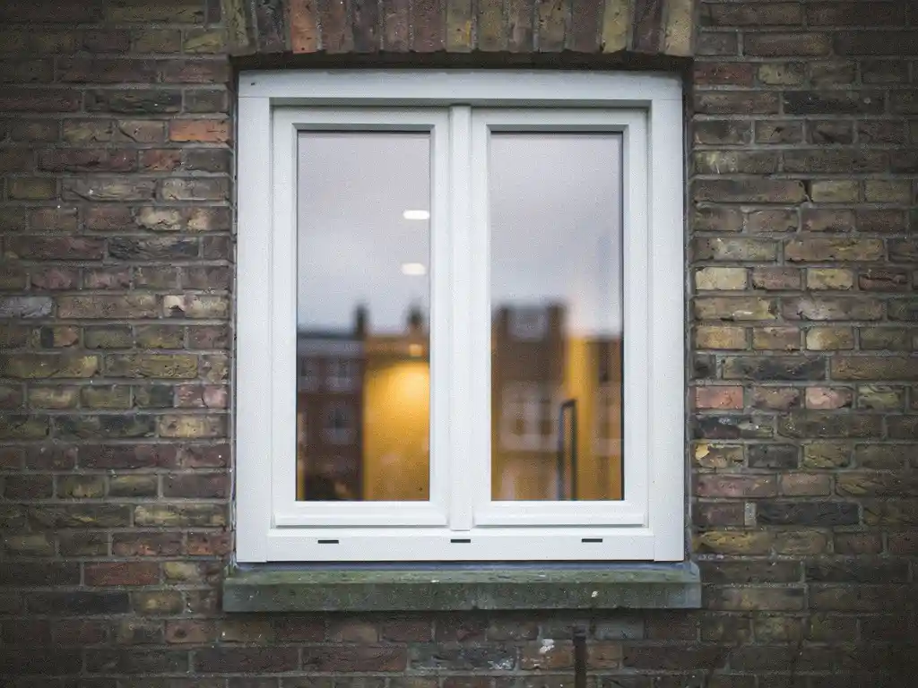 Wit uPVC raamkozijn in bakstenen gevel van Hollandse rijtjeswoning met warm binnenverlichting door driedubbel glas.
