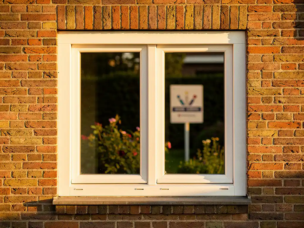 Nieuw wit PVC-raamkozijn in Nederlandse bakstenen gevel met dubbelglas, te koop-bord zichtbaar op de achtergrond.