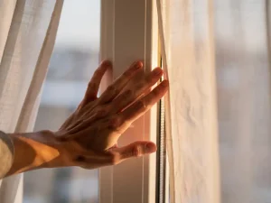 Huiseigenaar voelt koude tocht langs wit uPVC kozijn in woonkamer, licht opbollend gordijn op de achtergrond.