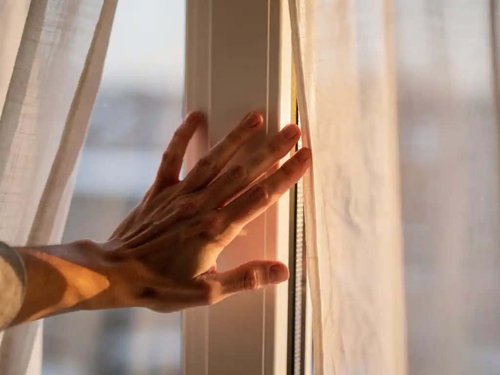Huiseigenaar voelt koude tocht langs wit uPVC kozijn in woonkamer, licht opbollend gordijn op de achtergrond.