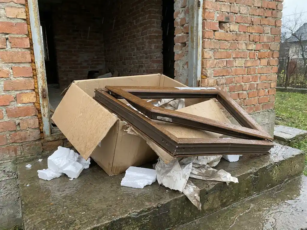 Vervormd plastic kozijn in open kartonnen doos op natte drempel, zichtbaar beschadigd bij aankomst, met verspreid verpakkingsschuim.