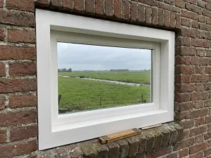Nieuw geplaatst wit kozijn op Friese boerderijgevel van rode baksteen, met meetlint en potlood op de vensterbank.