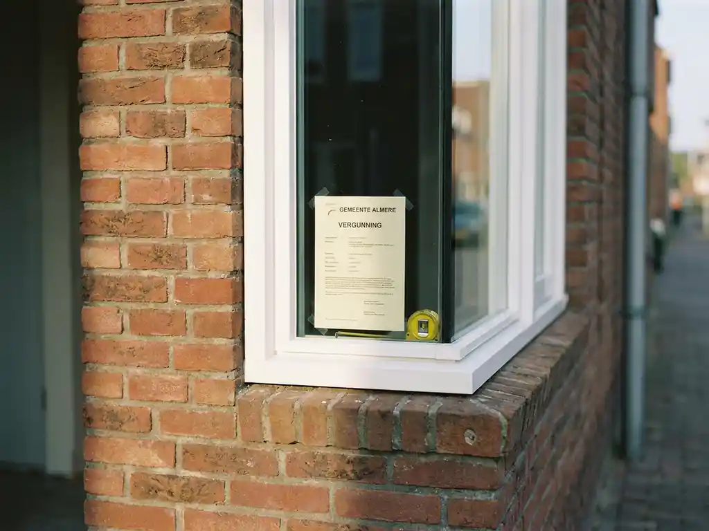 Nieuw wit PVC-kozijn in terracotta bakstenen gevel in Almere, met vergunningsdocument en meetlint op de vensterbank.