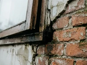 Loslatend kozijn in verweerde bakstenen muur met vochtschade, gebarsten pleisterwerk en donkere kieren aan Nederlandse woning.