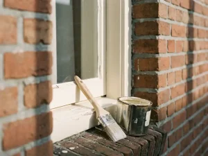 Vers geschilderd wit kozijn op een Nederlandse bakstenen gevel, met penseel en verfpot op de vensterbank in ochtendlicht.