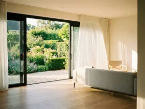 Wijd open schuifpui met uitzicht op een weelderige Nederlandse tuin, warm middaglicht valt binnen op lichte eiken vloer in moderne woonkamer.