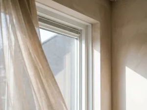 Wit uPVC raamkozijn met geïntegreerde ventilatiesleuf, gemonteerd in gepleisterde muur, doorzichtig gordijn beweegt zachtjes in de tocht.