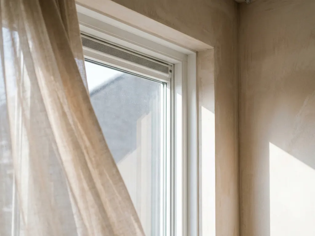 Wit uPVC raamkozijn met geïntegreerde ventilatiesleuf, gemonteerd in gepleisterde muur, doorzichtig gordijn beweegt zachtjes in de tocht.